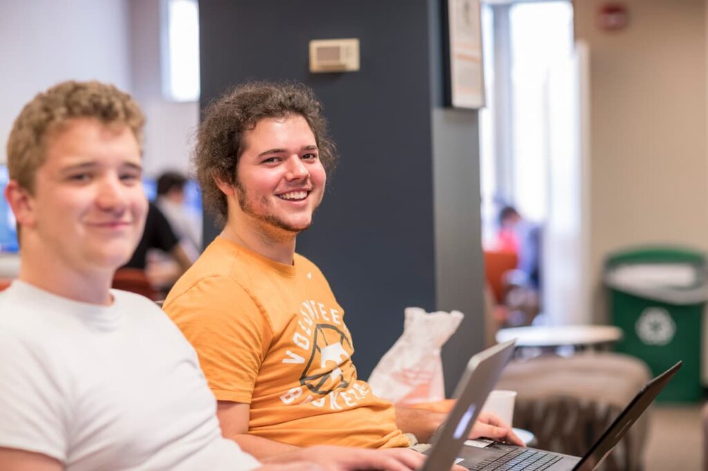 two male students facing camera with laptops