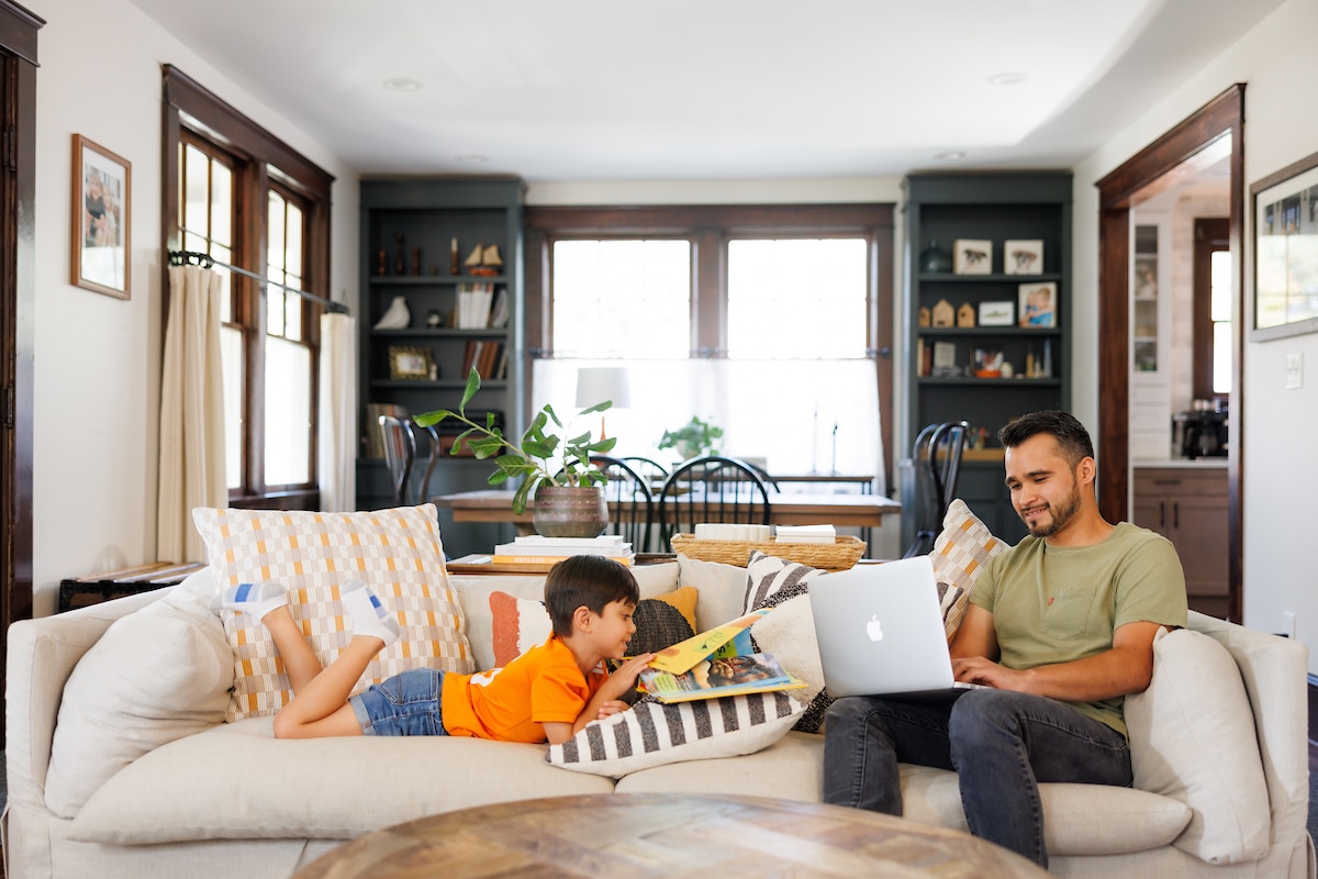 A father sits on a couch and works on a laptop as his son sits next to him reading a book during a Digital Learning produced lifestyle video/photoshoot inside a home in Fountain City by the Office of Communications and Marketing Department at the Uni